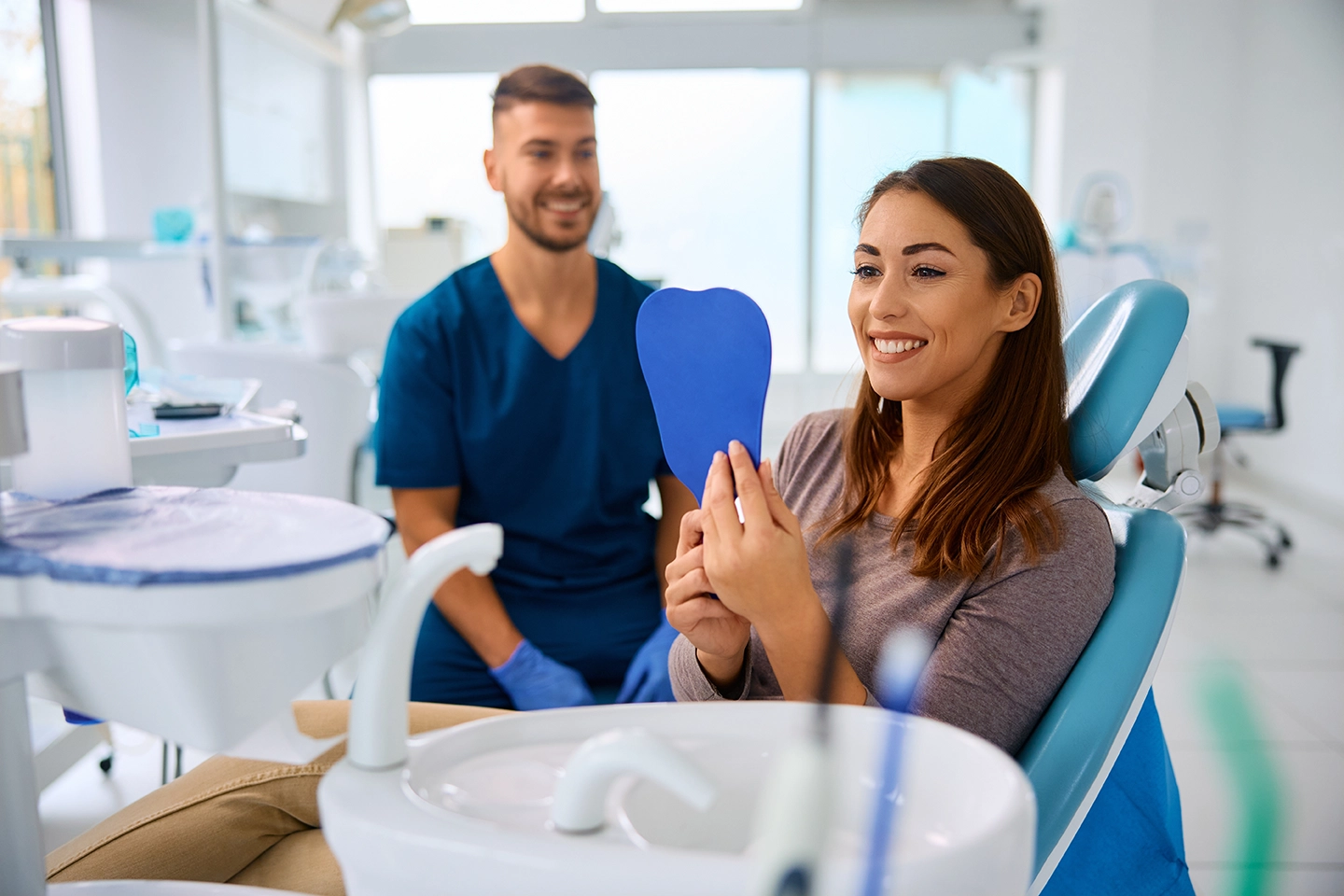 A woman looking at herself in the mirror while the dentist is smiling in the background.